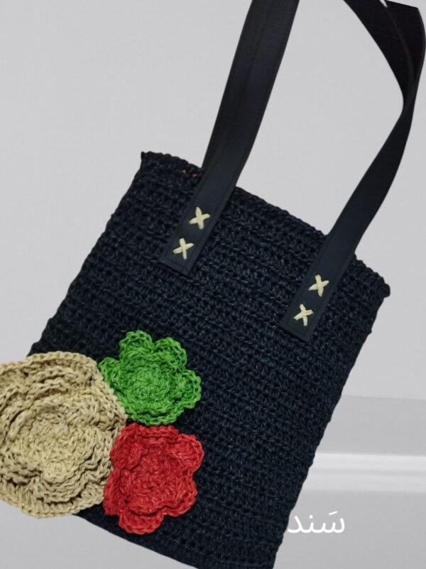 A black crocheted handbag with black handles, featuring three large crocheted flower appliqués in beige, green, and red on the lower left corner.