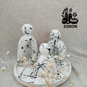 Three abstract, speckled white stone figures sit on a round tray, resembling a family. Black paint splatters decorate the figures. Dried flowers are in the foreground, and a logo with plants and SOKON is on the background fabric.