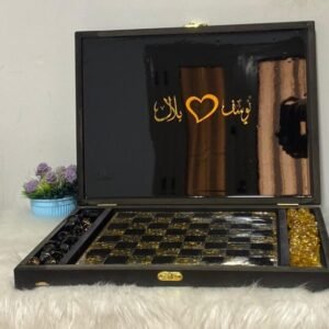 A black and gold chess and checkers set sits open on a fluffy surface, with gold Arabic calligraphy and a heart symbol on the inside of the lid. A small potted plant is in the background.