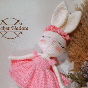 A crocheted doll wearing a pink dress with ruffled sleeves is shown from the shoulders down. The doll has white arms and legs. The logo Crochet.Hadota appears in the bottom left corner.