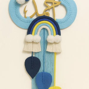 A handmade wall hanging shaped like a blue cloud with golden Arabic calligraphy, a rainbow in blue and yellow tones, white pom-poms, and three leaf-shaped tassels hanging down.
