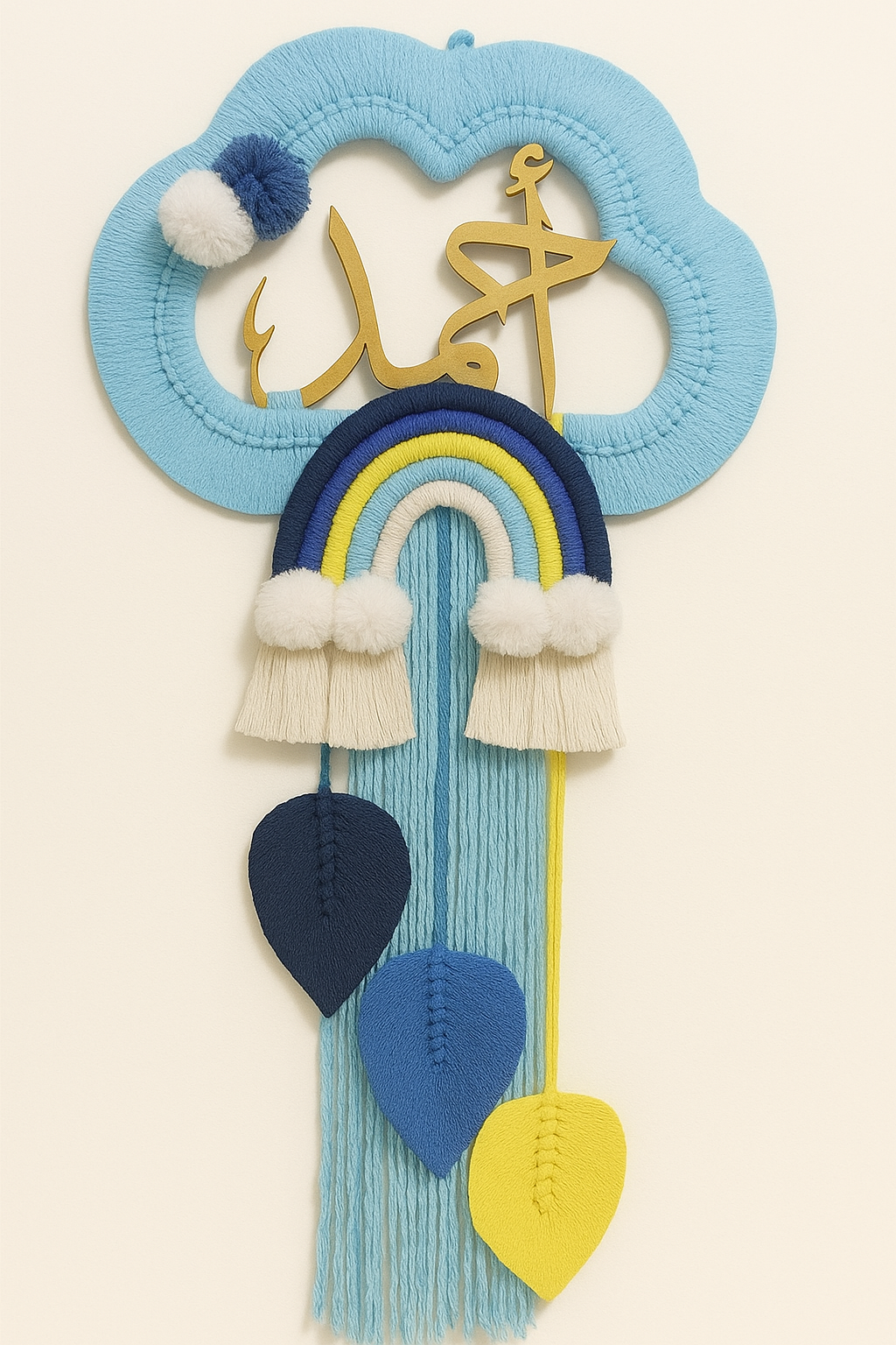 A handmade wall hanging shaped like a blue cloud with golden Arabic calligraphy, a rainbow in blue and yellow tones, white pom-poms, and three leaf-shaped tassels hanging down.