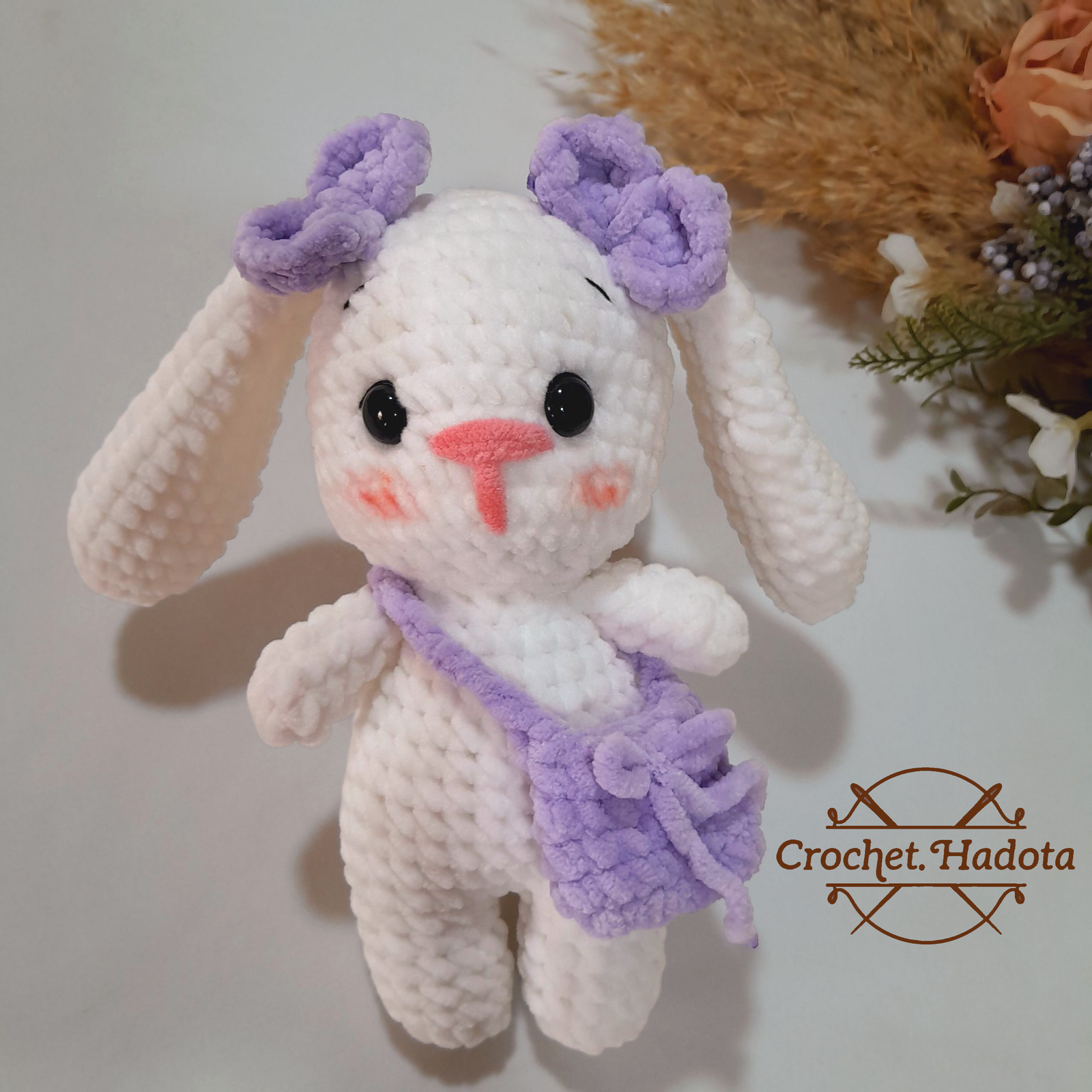 A crocheted rabbit wearing a bag is shown from the shoulders down. The rabbit has white arms and legs. The logo Crochet.Hadota appears in the bottom left corner.